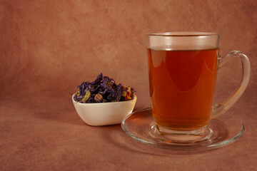 Glass cup of healthy, steaming mallow tea sits on a glass saucer, accompanied by dried mallow...