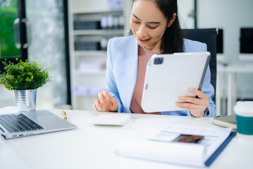 Asian businesswoman in blue blazer using tablet at modern office desk with laptop, coffee