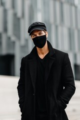 A stylish man wearing a black coat and cap, with a black mask mockup, standing outdoors against a modern architectural background