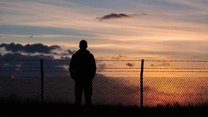 Man stands silhouetted against a fiery sunset, looking over a barbed wire fence - Powered by Adobe