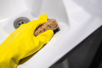 Cleaning the bathroom sink, she is polishing the faucet using a sponge, protective yellow gloves and spray detergent.