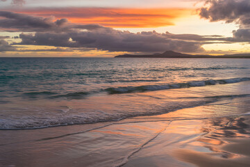 Fototapeta premium Ocean Sunset Serenity: A picturesque coastal panorama unfolds as the sun dips below the horizon, casting a warm glow over gentle waves washing onto the sandy shore and clouds.