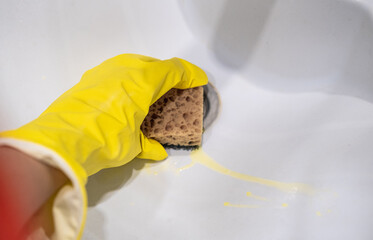 Cleaning the bathroom sink, she is polishing the faucet using a sponge, protective yellow gloves and spray detergent.