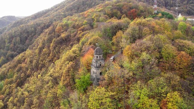 A cinematic drone shot slowly orbits Monumentul Eroilor in Targu Ocna, Romania. The structure is surrounded by a forest of trees in peak autumn color, with a monastery above.