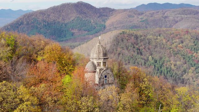 A cinematic aerial shot pulls back to reveal the Heroes' Monument in Targu Ocna, Romania, standing on a hilltop above a forest of vibrant autumn colors and the town below.