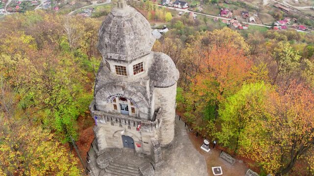 A cinematic aerial shot slowly orbits the Heroes' Monument in Targu Ocna, Romania. The stone tower stands on a hilltop surrounded by a forest of vibrant autumn colors.