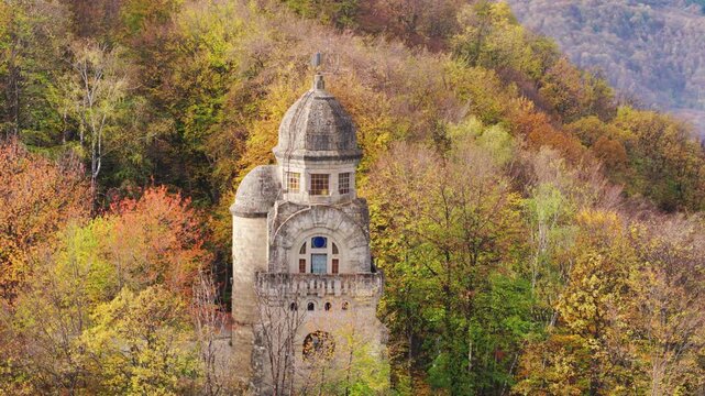 A cinematic close-up aerial shot slowly orbits the stone tower of the Heroes' Monument in Targu Ocna, Romania, surrounded by the vibrant colors of an autumn forest.