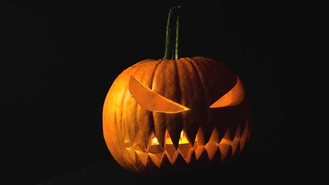 Close up video orbiting around a freshly carved Halloween orange pumpking with an evil jack o lantern face carved into it. The pumpkin has candles burning inside. In front of black studio background.