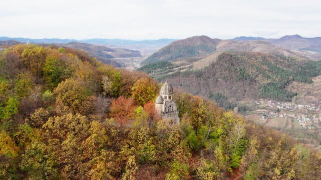 A cinematic aerial shot slowly orbits a historic Monumentul Eroilor in Targu Ocna, Romania, a war memorial nestled among a forest of trees in peak autumn color.