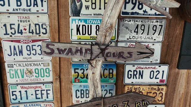 Close-up view of a rustic wooden sign pointing to swamp tours, placed in front of a wall decorated with colorful vintage American license plates in Louisiana, USA.