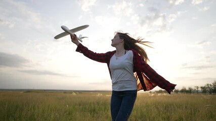 Woman launching toy plane at sunset across wide field of tall grass and open sky, girl extends arm with toy plane showing freedom and joy in nature, casual outfit and windblown hair and golden light