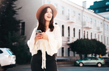 Fototapeta premium Young beautiful smiling woman in trendy summer clothes. Carefree female posing in street in sunny day. Positive model holds smartphone, looks at cellphone screen, uses mobile apps, in hat
