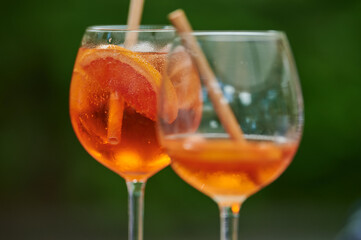 A close-up of two cocktail glasses, each filled with an orange-hued drink and garnished with a slice of citrus fruit, against a blurred green background