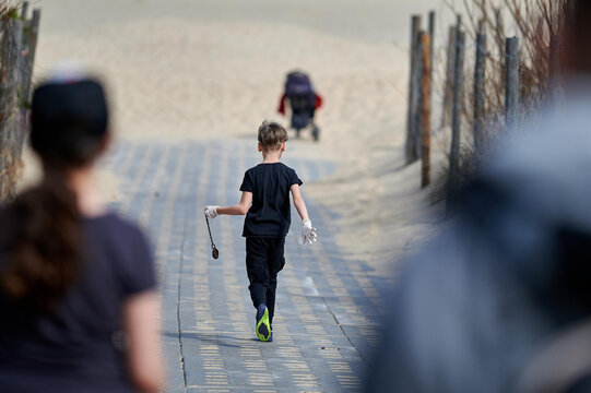 A young boy runs ahead on a sandy path holding a small object in hand, participating in a cleanup activity at the beach, while wearing protective gloves and casual black clothing