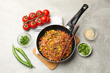 Delicious fried rice in frying pan, chopsticks and ingredients on grey textured table, flat lay