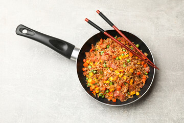 Delicious fried rice with vegetables in frying pan and chopsticks on grey textured table, top view