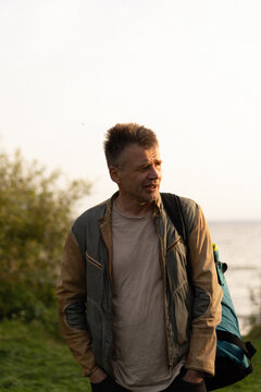 Man standing outside by the water, looking thoughtful with a backpack
