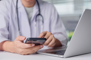 Doctor uses smartphone next to laptop in clinic office, telemedicine ehealth patient consult message schedule record management secure communication concept