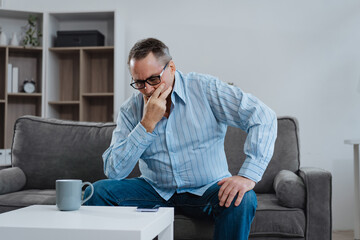 A middle-aged Caucasian man sits on a sofa, appearing stressed with a headache and...