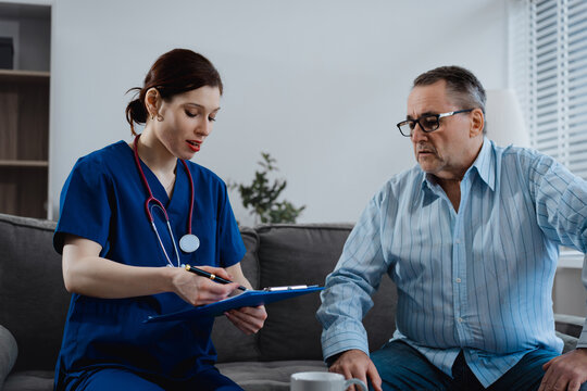A young Caucasian female nurse offers medical consultation and healthcare support to an elderly Caucasian male patient sitting on a sofa, focusing on examination, treatment,and compassionate patient