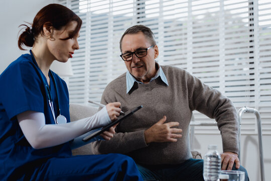 A young Caucasian nurse provides medical consultation to an elderly Caucasian man sitting on a sofa, addressing chronic conditions like obesity, hypertension, diabetes,heart disease, dementia, joint