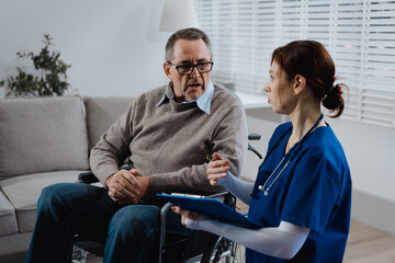 An elderly male patient in a wheelchair discusses neck pain with a young nurse taking notes in a...