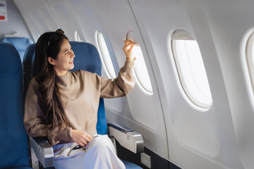 Fototapeta premium An Asian woman sits comfortably on an airplane, holding her smartphone smiling while looking out the window, representing modern travel,connectivity, leisure,the joy of experiencing a flight journey