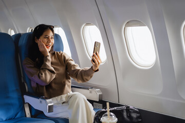 Fototapeta premium An Asian woman sits comfortably on an airplane, holding her smartphone smiling while looking out the window, representing modern travel,connectivity, leisure,the joy of experiencing a flight journey