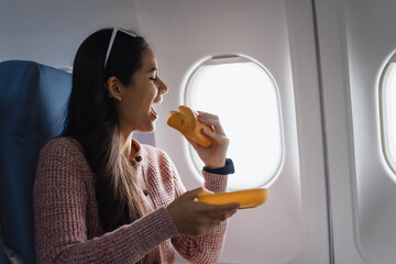 Happy Asian woman enjoying a sandwich on an airplane. Relishing her in-flight meal, she savors every bite, making the travel experience more enjoyable and memorable before reaching her destination