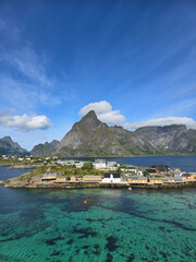 Breathtaking panorama of Sakrisoy in Lofoten, Norway, showcasing colorful houses along the shoreline against vivid turquoise waters and dramatic mountain backdrops, under a clear blue sky.