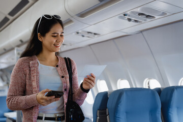 An Asian woman sits near the airplane window, smiling warmly while wearing a casual pink sweater...