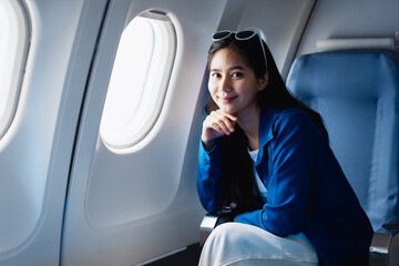 Fototapeta premium A cute Asian woman enjoys a fun travel experience on a plane, smiling happily while sitting by the window, reflecting the relaxation, freedom and excitement of her travel during the trip