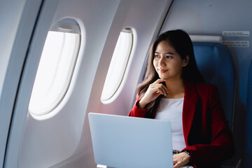 Fototapeta premium A lovely Asian businesswoman sits at the airplane window, working with a laptop and holding a smartphone confidently,demonstrating her efficiency,professionalism and modern travel lifestyle on board