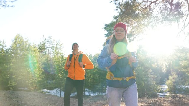 Couple hiking through forest trail with backpack on pine under sunlight lens flare outdoor nature walk winter snow patch man in orange jacket woman in red beanie following enjoying outdoor adventure