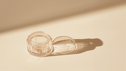 Transparent cosmetic jar and lid resting on beige tabletop, casting warm directional shadows