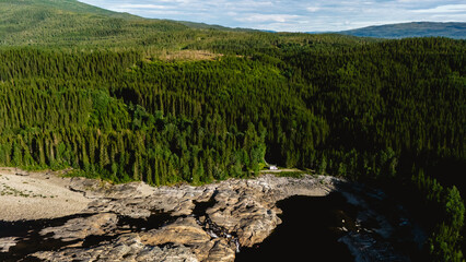 Vibrant green forests cover the landscape, contrasting with the rocky edges of a tranquil river. A small cabin peeks through the trees, inviting exploration in Norways natural beauty.