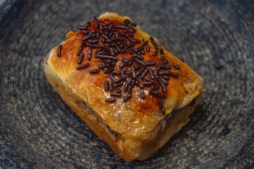 Golden puff pastry slice with flaky crust and soft filling, topped with chocolate sprinkles on dark plate ideal for showcasing baked texture, sweet topping, and dessert appeal.