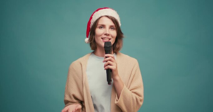 Friendly Hispanic woman in red santa hat with mic makes Christmas announcement and happy smiling on blue background