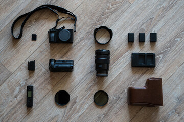 Flat lay of photography equipment on wooden floor: camera, lens, batteries, charger, filters, grip, remote and leather case. Minimalistic professional photorapher set