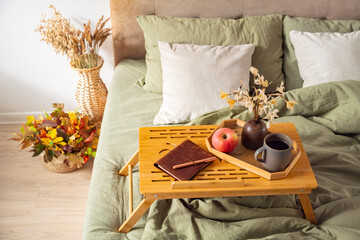 Cozy bed scene featuring wooden breakfast tray with coffee, apple and notebook on green linen...
