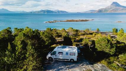 A serene coastal view in Bognes, Lofoten, where a camper van is parked among lush greenery, overlooking calm waters and distant mountains under a clear blue sky during the day. © Fokke Baarssen