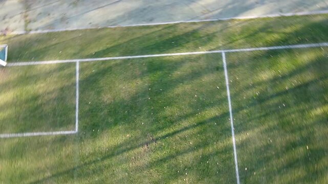 Aerial view of a football pitch corner and goal box with long evening shadows on green grass. Minimal, geometric sports field lines. No people.