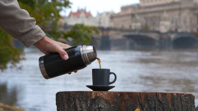 Hot drink from a thermos on the river bank. Tourist pours coffee or tea from a thermos into a cup placed on a tree stump on the banks of the river in prague, with a view of the bridges