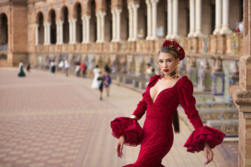 Young, beautiful, blonde woman in red flamenco dress, flowers in her hair and gold jewelry, performing flamenco dance poses in the square of Spain in Seville. Dance concept, art, typical Spanish.