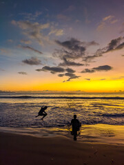 Golden sunset paints the sky while two kids create playful silhouettes along the gentle waves of Kuta Beach, capturing carefree joy as colorful reflections shimmer across the wet shoreline.