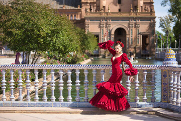 Naklejka premium Young, beautiful, blonde woman in red flamenco dress, flowers in her hair and gold jewelry, performing flamenco poses leaning on a tiled railing in the square of Spain in Seville. Dance concept, art.