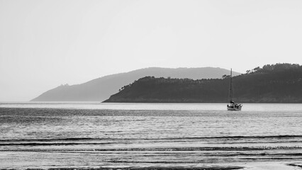 Black and white photograph of a sailboat in the Atlantic with Cape Finisterre and its lighthouse in the background.