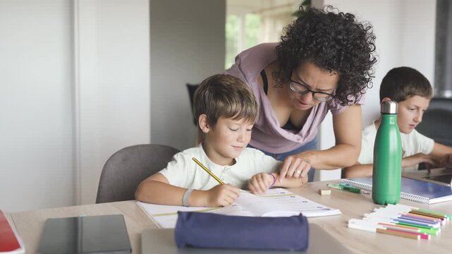 Mother helping young son with homework while older brother studies on laptop at home