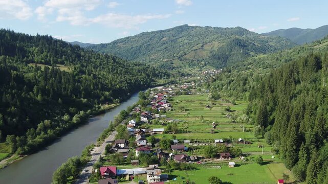 A static, high-angle aerial view of a rural village in a Romanian mountain valley. Houses are scattered along a river and road, surrounded by lush green summer forests and fields. Brosteni, Suceava.