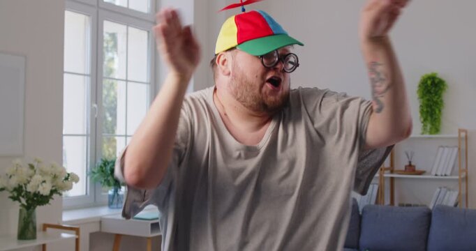 Chubby man dancing goofy at home livingroom. With glasses and a propeller cap, he shows funny, silly moves in an apartment, radiating happy vibes and body positivity. Playful dance concept.